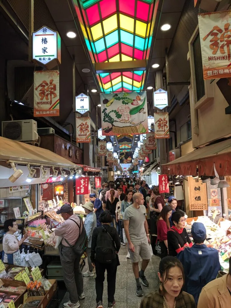 A bustling scene of shoppers in the Nishiki Market in Kyoto, with colorful overhead lights and signs.