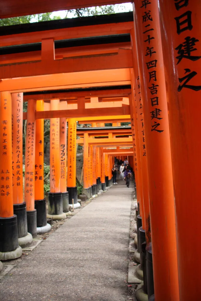 A pathway lined with red torii gates at Fushimi Inari Taisha, featuring inscriptions along the gates and visitors walking through.