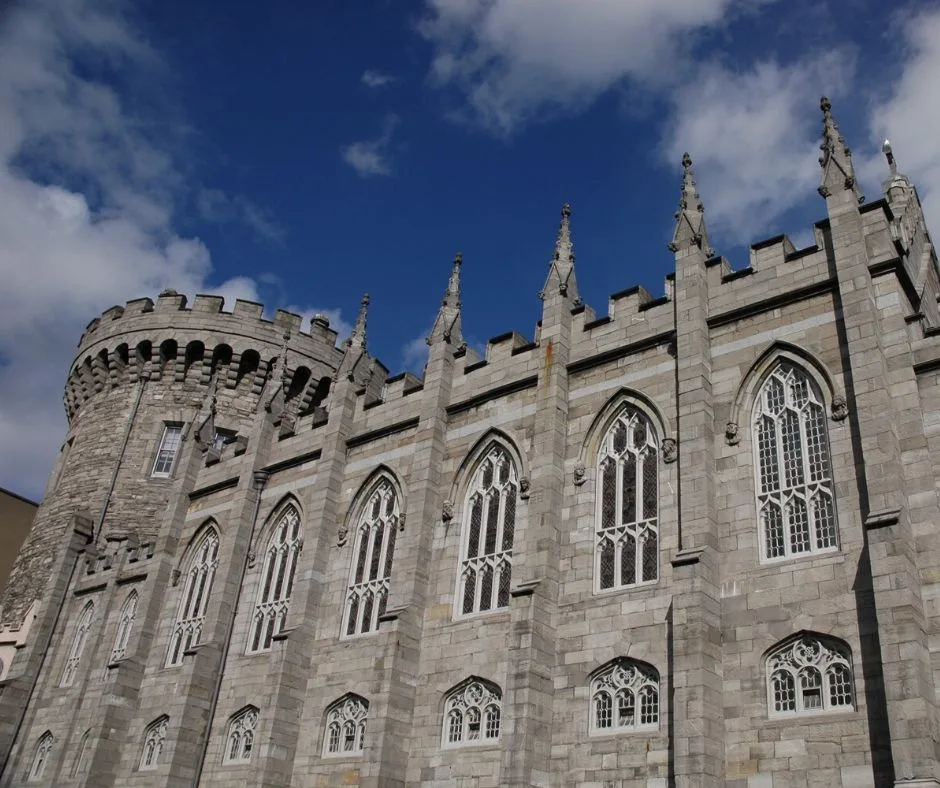 View of Dublin Castle, showcasing its Georgian architecture with symmetrical facades, a central courtyard, and the medieval Record Tower. The building features classic arches, columns, and decorative stonework, set against a cloudy sky.