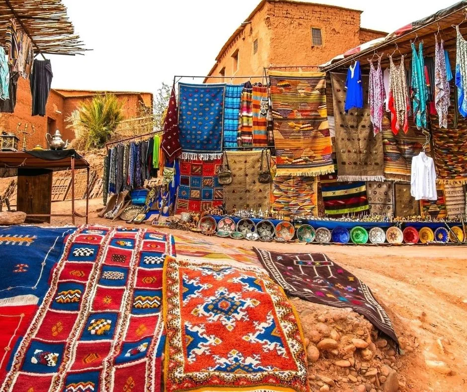 A vibrant market in Marrakech, Morocco, with colorful rugs, textiles, and pottery displayed on the ground and hanging from stalls.