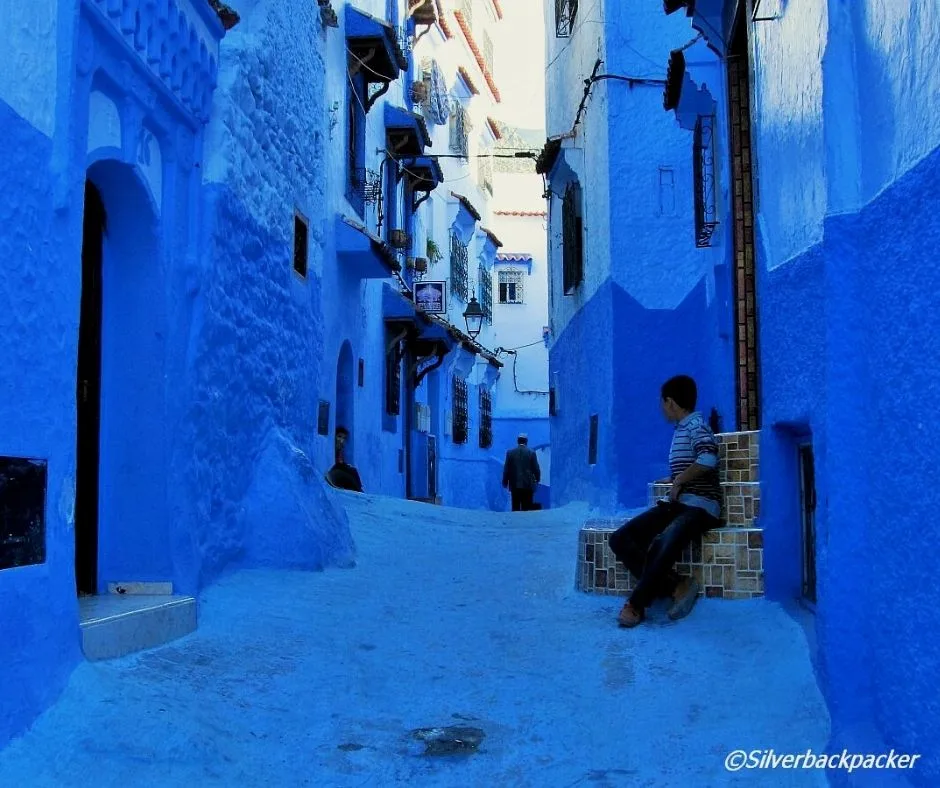 A narrow blue street in Chefchaouen, Morocco, with a child sitting on a tiled bench.