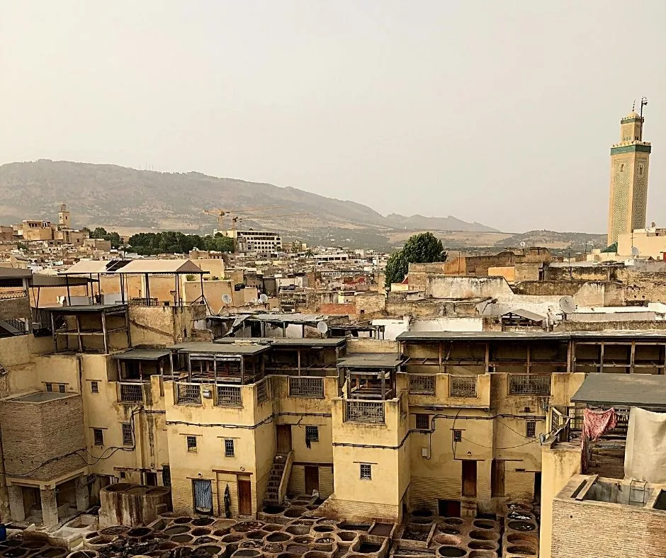 A view of the cityscape of Fes, Morocco, with rooftops, a mosque minaret, and the surrounding mountains in the distance.
