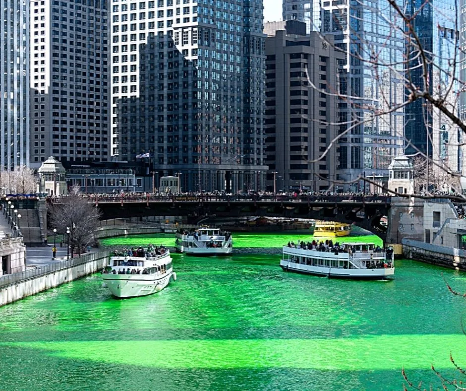 Boats cruising along the Chicago River, dyed green for St. Patrick's Day, surrounded by skyscrapers.